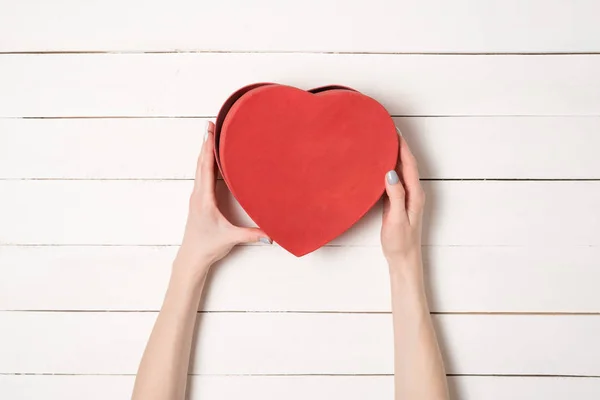 Las manos femeninas sostienen una caja en forma de corazón rojo contra el fondo de una mesa de madera blanca — Foto de Stock