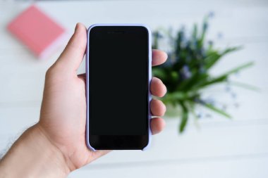 Akıllı telefonlu erkek eli. Siyah boş ekran. Arka planda dizüstü bilgisayar ve çiçekleriçeren tablo