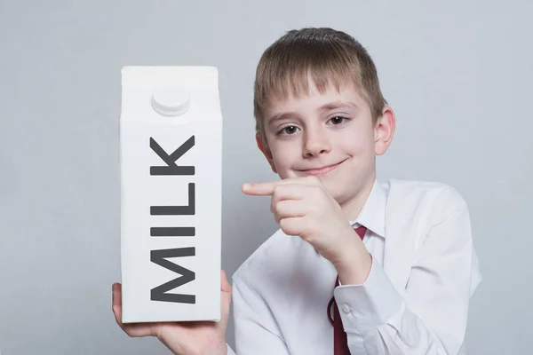 Il bambino biondo tiene e mostra l'indice su una grande confezione di latte di cartone bianco. Camicia bianca e cravatta rossa. Sfondo chiaro — Foto Stock