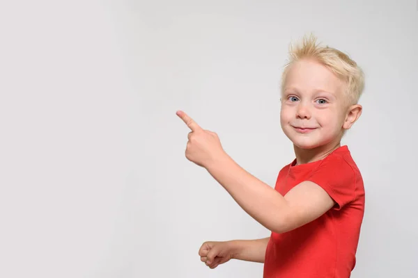 빨간 티셔츠에 작은 금발의 소년은 자신의 손가락을 가리킵니다. 텍스트의 공백입니다. 광고를 위한 장소. 흰색 배경 — 스톡 사진