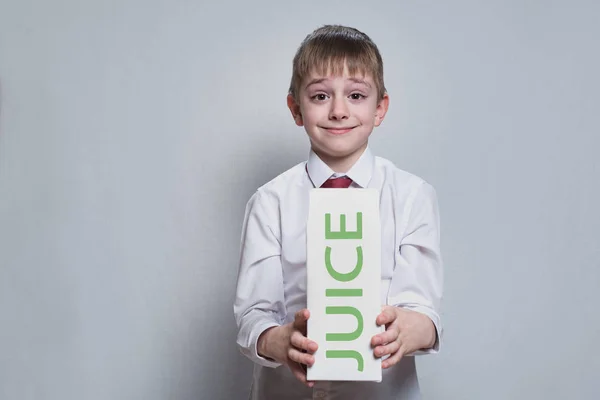 Piccolo ragazzo biondo tiene e mostra un grande pacchetto di succo di cartone bianco. Camicia bianca e cravatta rossa. Sfondo chiaro — Foto Stock