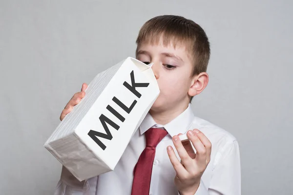 Il bambino beve da un grande pacchetto di latte bianco. Camicia bianca e cravatta rossa. Sfondo chiaro — Foto Stock