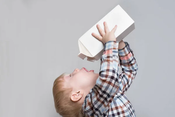 Der kleine blonde Junge trinkt aus einem großen weißen Päckchen. heller Hintergrund — Stockfoto