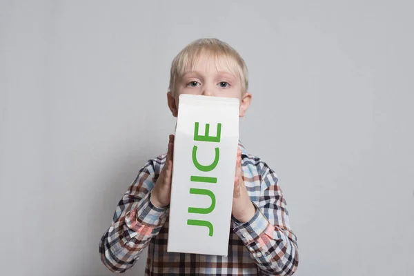 Il ragazzino biondo ha in mano un grosso pacchetto di succo di cartone bianco. Sfondo chiaro . — Foto Stock