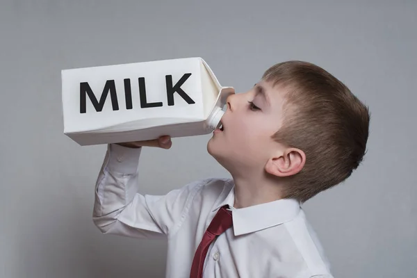 Ένα μικρό αγόρι πίνει από ένα μεγάλο λευκό πακέτο γάλακτος. Λευκό πουκάμισο και κόκκινη γραβάτα. Φως φόντο — Φωτογραφία Αρχείου