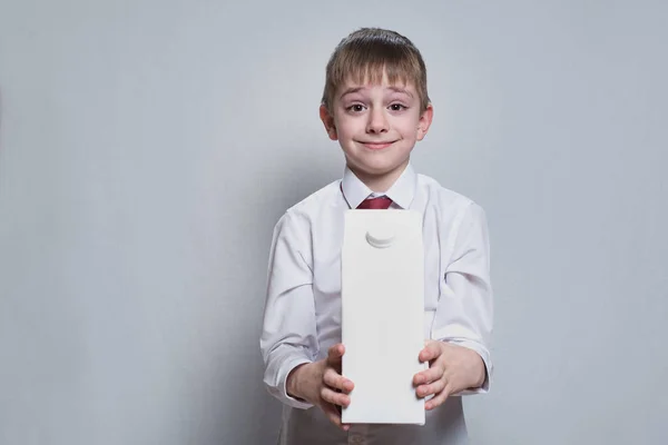 Kleine blonde jongen houdt en toont een grote witte kartonverpakking. Wit overhemd en rode stropdas. Lichte achtergrond — Stockfoto