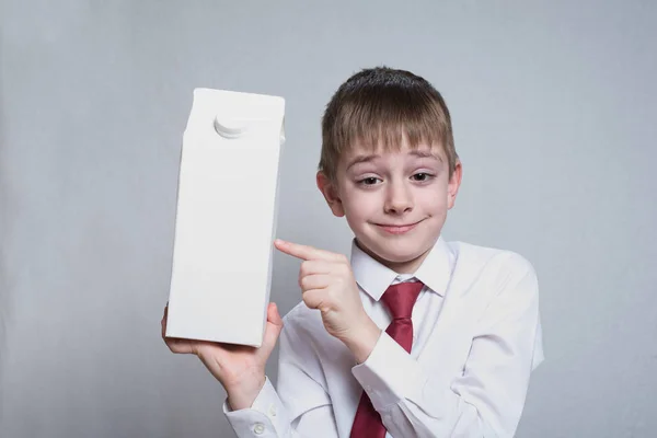 Kleine blonde jongen houdt en toont de wijsvinger op een groot wit karton pakket. Wit overhemd en rode stropdas. Lichte achtergrond — Stockfoto