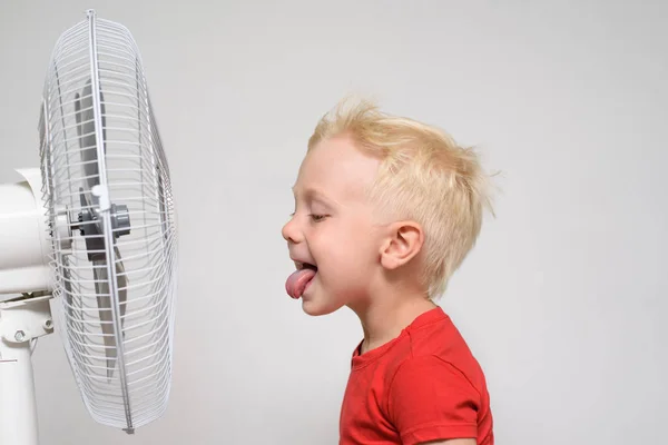 Chico rubio divertido con una camiseta roja cerca del ventilador con su lengua sobresaliendo. Disfruta del aire fresco. Concepto de verano —  Fotos de Stock