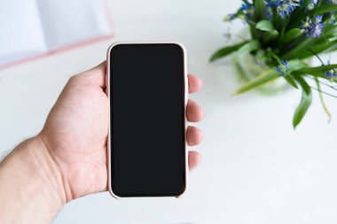 Akıllı telefonlu erkek eli. Siyah boş ekran. Arka planda dizüstü bilgisayar ve çiçekleriçeren tablo