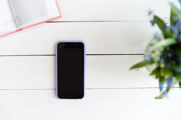 Smartphone con pantalla vacía en negro. Cuaderno, lupa y flores en la mesa — Foto de Stock