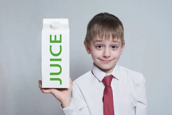 little blond boy holds and shows a big white carton juice package. White shirt and red tie. Light background