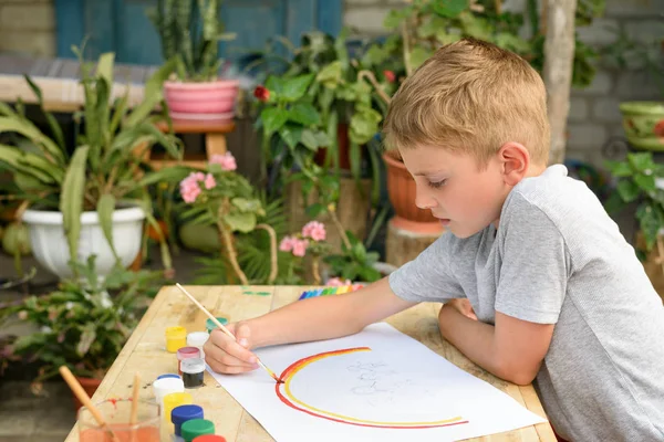 Cute boy draws rainbow. Open air. Garden in the background. Creative concept.