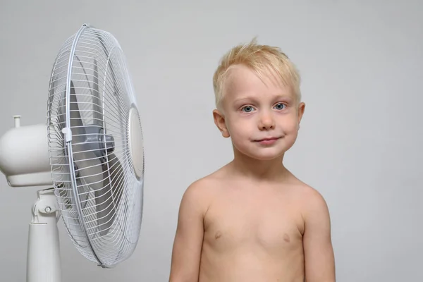 Niño rubio bastante sonriente con el torso desnudo se encuentra cerca de un ventilador. Concepto de verano —  Fotos de Stock