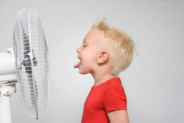 Funny blond boy in a red T-shirt near the fan with his tongue sticking out. Enjoy cool air. Summer concept