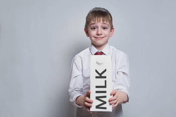 Kleine blonde jongen houdt en toont een grote witte kartonnen melk pakket. Wit overhemd en rode stropdas. Lichte achtergrond — Stockfoto
