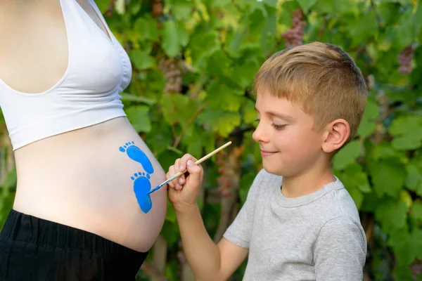 El niño dibuja con un cepillo en el vientre de la huella de su madre embarazada. Concepto de embarazada. Primer plano —  Fotos de Stock