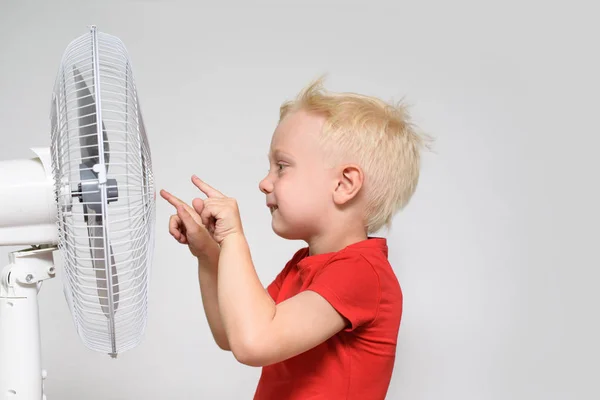 El niño rubio con una camiseta roja toca el ventilador con el dedo. Concepto de verano —  Fotos de Stock