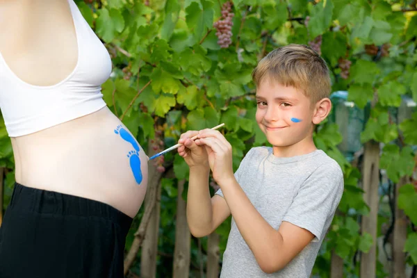 Szczęśliwy chłopiec rysuje z pędzlem na brzuchu jego ciąży mama dziecka ślad. Koncepcja w ciąży. Zbliżenie — Zdjęcie stockowe