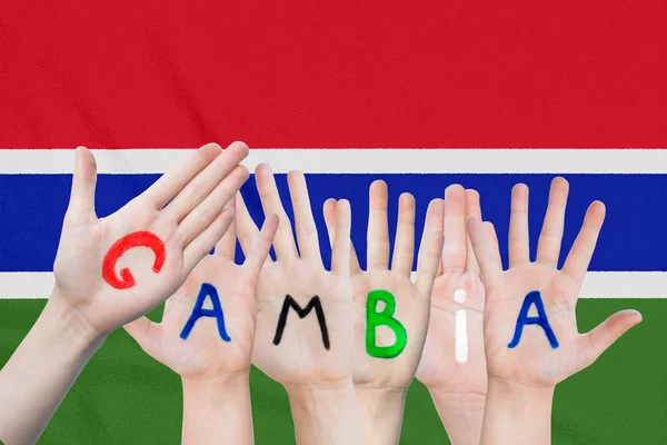 Inscrição Gâmbia nas mãos das crianças contra o fundo de uma bandeira ondulante da Gâmbia — Fotografia de Stock