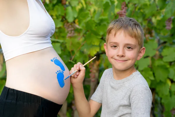 Happy little boy dibuja con un cepillo en el vientre de la huella de su madre embarazada. Concepto de embarazada. Primer plano —  Fotos de Stock