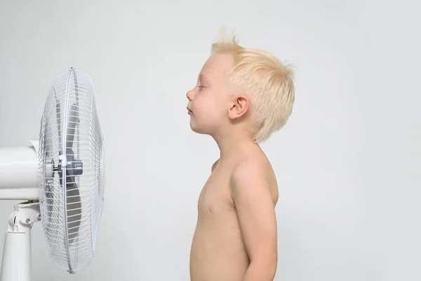 En liten blond gutt med naken overkropp og lukkede øyne står nær en vifte. Sommerkonsept – stockfoto