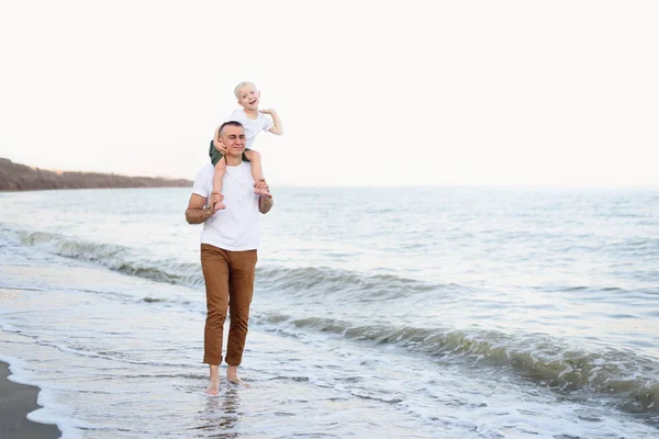 Padre con un hijo rubio sobre sus hombros están caminando a lo largo de la orilla del mar. Vacaciones familiares — Foto de Stock