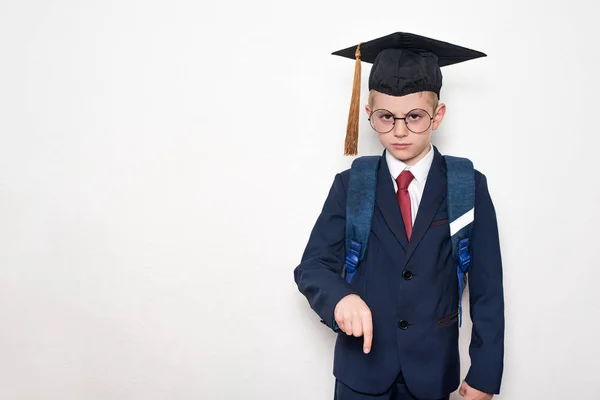 Takım elbiseli, gözlüklü ve akademik şapkalı ciddi bir öğrenci aşağıyı gösteriyor. Okul konsepti. Beyaz arka plan. — Stok fotoğraf