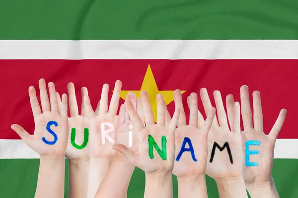 Inscrição Suriname nas mãos das crianças contra o fundo de uma bandeira ondulante do Suriname — Fotografia de Stock
