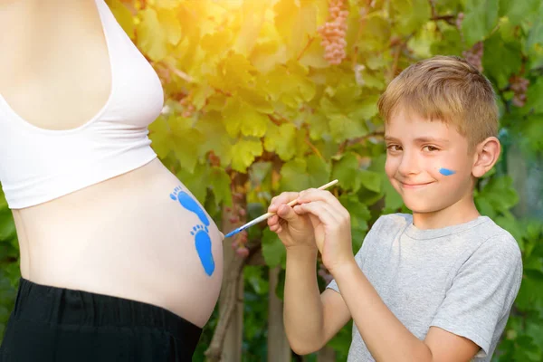 Joyeux petit garçon dessine avec un pinceau sur le ventre de l'empreinte de pas de bébé de sa mère enceinte. Concept de grossesse. Gros plan — Photo