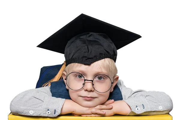 Portret van een schattige blonde jongen in grote glazen, academische hoed en een rugzak. Isoleren. — Stockfoto