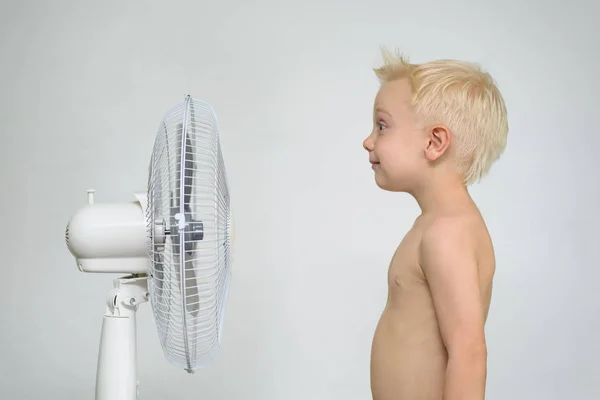 Un niño rubio con el torso desnudo está cerca de un ventilador. Concepto de verano — Foto de Stock