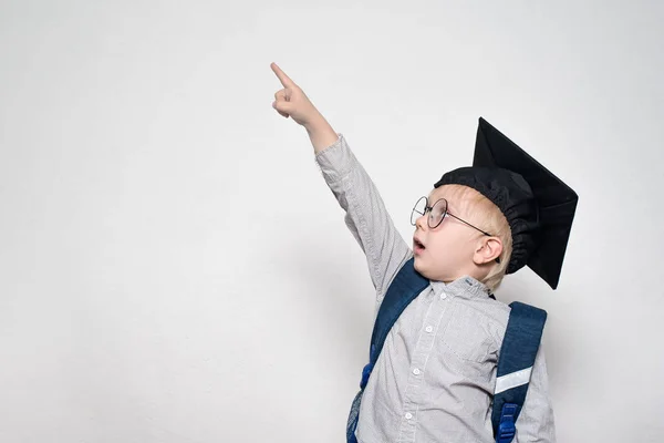 Uno scolaro sorpreso con un completo, occhiali e un cappello accademico punta il dito verso l'alto. Concetto scolastico. Fondo bianco . — Foto Stock