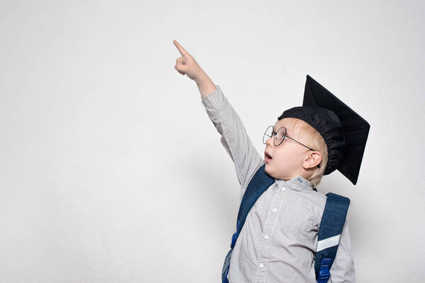 Surprised schoolboy in a suit, glasses and an academic hat points his finger up. School concept. White background.