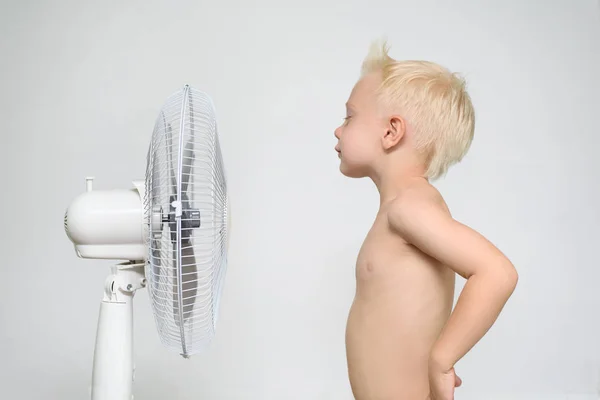 Niño rubio con el torso desnudo y los ojos cerrados se encuentra cerca de un ventilador. Concepto de verano —  Fotos de Stock