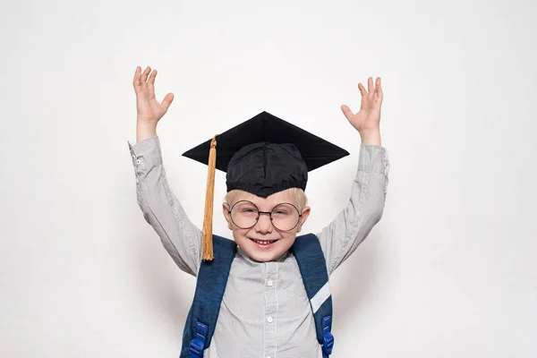 Portrait d'un garçon blond joyeux dans de grandes lunettes, chapeau académique et un sac à dos. Les mains en l'air. Fond blanc . — Photo