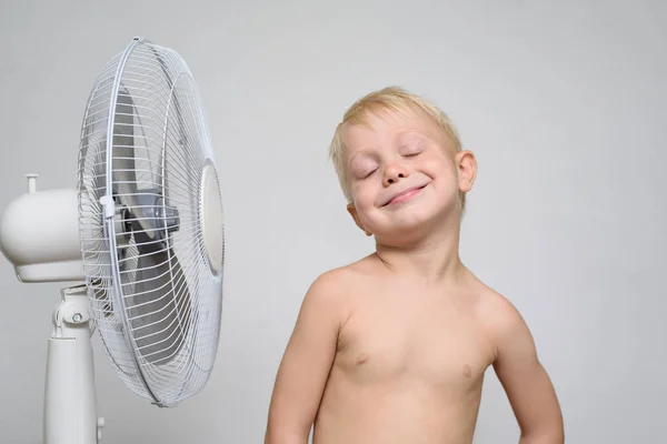Niño rubio bastante sonriente con el torso desnudo y los ojos cerrados se encuentra cerca de un ventilador. Concepto de verano —  Fotos de Stock