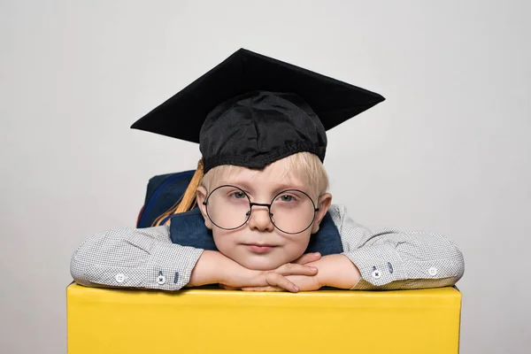 Portret cute blond chłopca w dużych okularach, kapelusz akademicki i plecak. Białe tło. — Zdjęcie stockowe