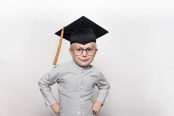 Portrait d'un mignon garçon blond dans de grandes lunettes et un chapeau académique. Fond blanc . — Photo