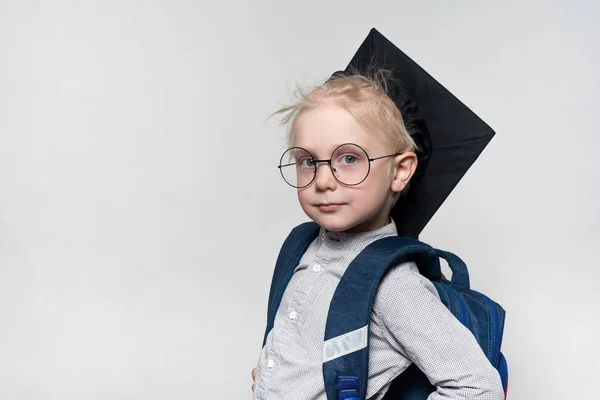 Portrait d'un garçon blond en lunettes, un chapeau académique et un cartable sur fond blanc. Concept scolaire — Photo