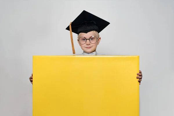 Garçon blond dans un chapeau académique et des lunettes tient une grande boîte. Fond blanc. Concept scolaire — Photo