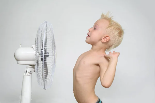 Menino loiro surpreso com tronco nu fica perto de um ventilador. Conceito de verão — Fotografia de Stock