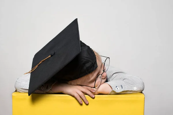 Garçon blond avec un chapeau académique et des lunettes endormies sur la table. Fond blanc. Concept scolaire — Photo