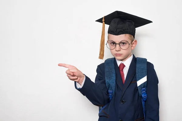 Un écolier sérieux en costume, lunettes et chapeau scolaire pointe du doigt. Concept de l'école. Fond blanc . — Photo