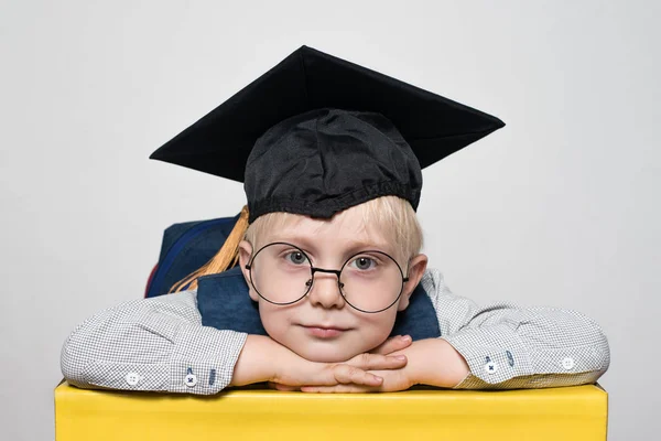 Portrait d'un mignon garçon blond en grands verres, chapeau académique et un sac à dos. Fond blanc . — Photo