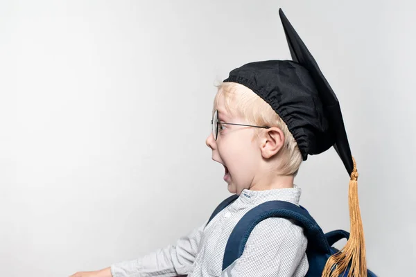Gözlük bir sürpriz sarışın çocuk portre, akademik bir şapka ve beyaz bir arka planda bir okul çantası. Okul konsepti — Stok fotoğraf