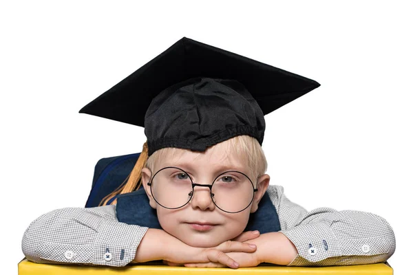 Portret van een schattige blonde jongen in grote glazen, academische hoed en een rugzak. Isoleren. — Stockfoto