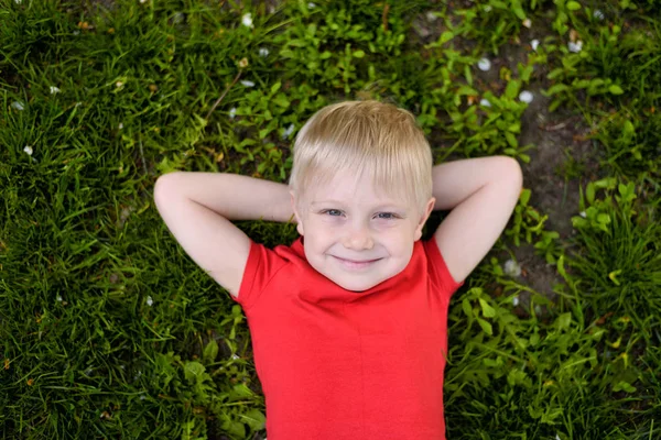 Portret uśmiechniętego blond chłopca leżącego na trawie. Ręce za głową. Odpoczynek na łonie natury — Zdjęcie stockowe