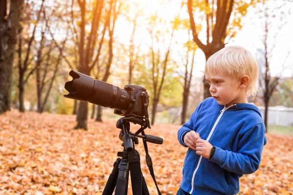 Menino loiro dispara com uma grande câmera SLR em um tripé. Sessão de fotos no parque de outono — Fotografia de Stock