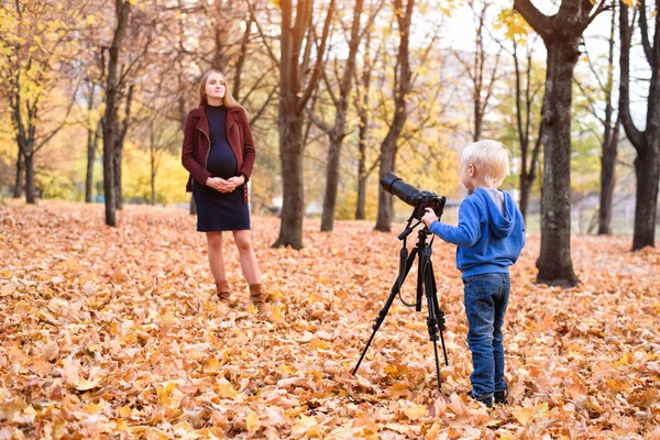 Malý blonďák s velkou reflexní kamerou na trojnožce. Fotografie těhotné ženy. Rodinná fotografická relace — Stock fotografie