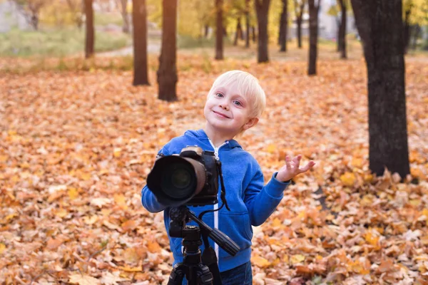 Portrét malého blonďáku s velkou DSLR kamerou na trojnožce. Fotografická relace v podzimním parku — Stock fotografie